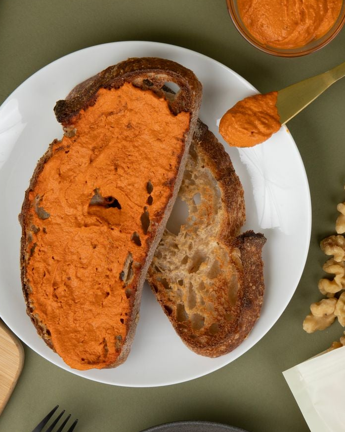Toast with romesco sauce on a white plate, accompanied by a jar of the same spread and walnuts on a green background.