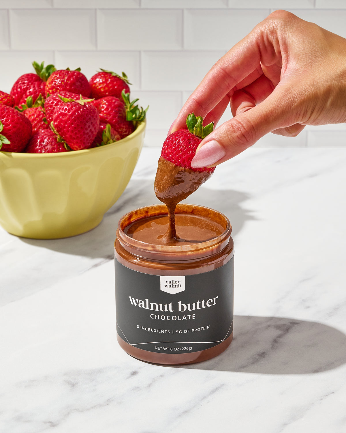 A hand dips a strawberry into a jar of Chocolate Walnut Butter, with a bowl of fresh strawberries in the background on a white marble countertop.